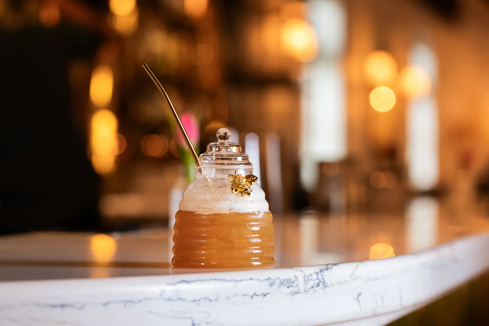 An amber drink with a foam top in a lidded glass jar shaped like a beehive, with a small gold bee and a gold straw, on a marble counter, warm lights blurred behind