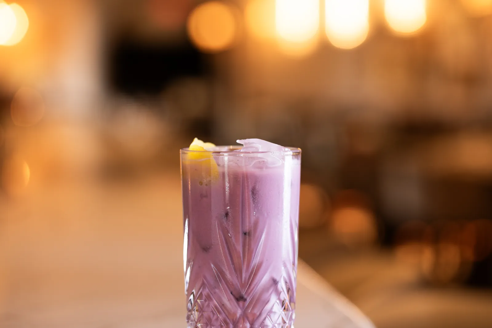 A pale purple drink with ice and a lemon twist in a cut-crystal glass, warm lights blurred behind