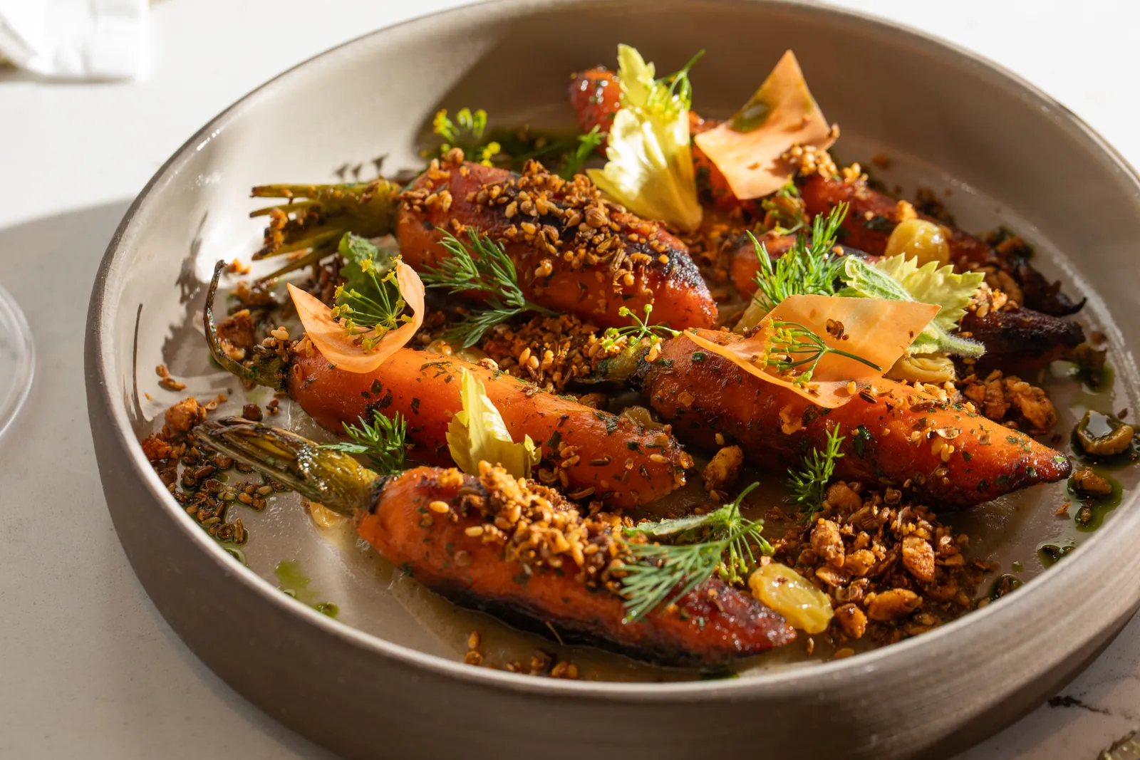 Whole roasted carrots under a golden crumble, with dill and thin, pale-orange shavings, in a grey bowl