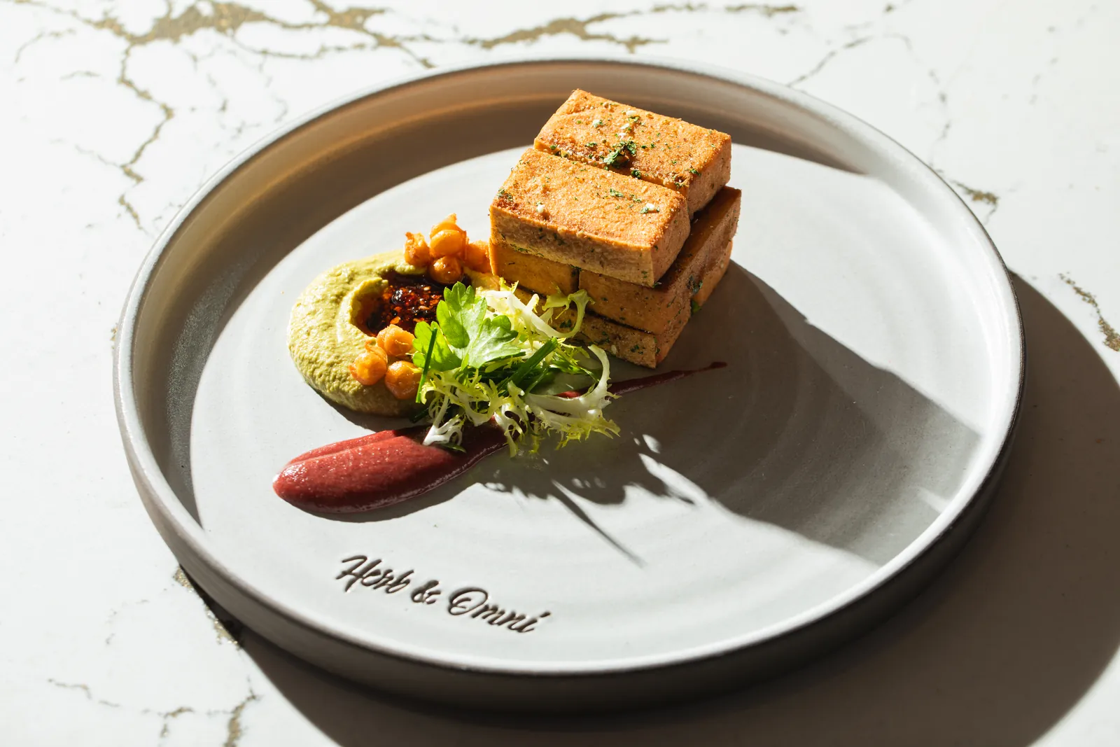 Three stacked golden-brown blocks with green purée, crisp chickpeas and frisée on a grey plate with “Herb & Omni” written in script near the rim
