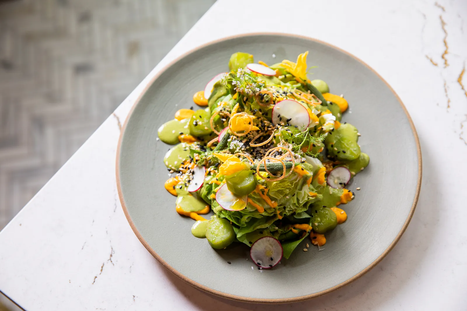 A lettuce salad with sliced radish, glossy green vegetable pieces, pools of green sauce, dots of orange sauce, yellow flowers and sesame on a grey plate