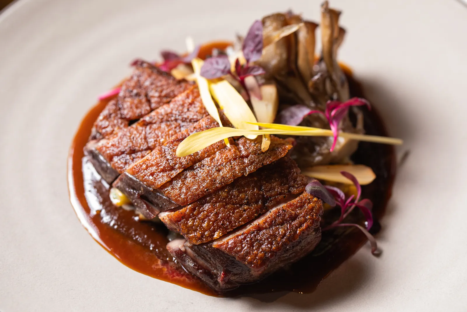 Slices of seared meat with a crisp, scored skin over a dark sauce, topped with frilled mushrooms, purple leaves and pale yellow slivers
