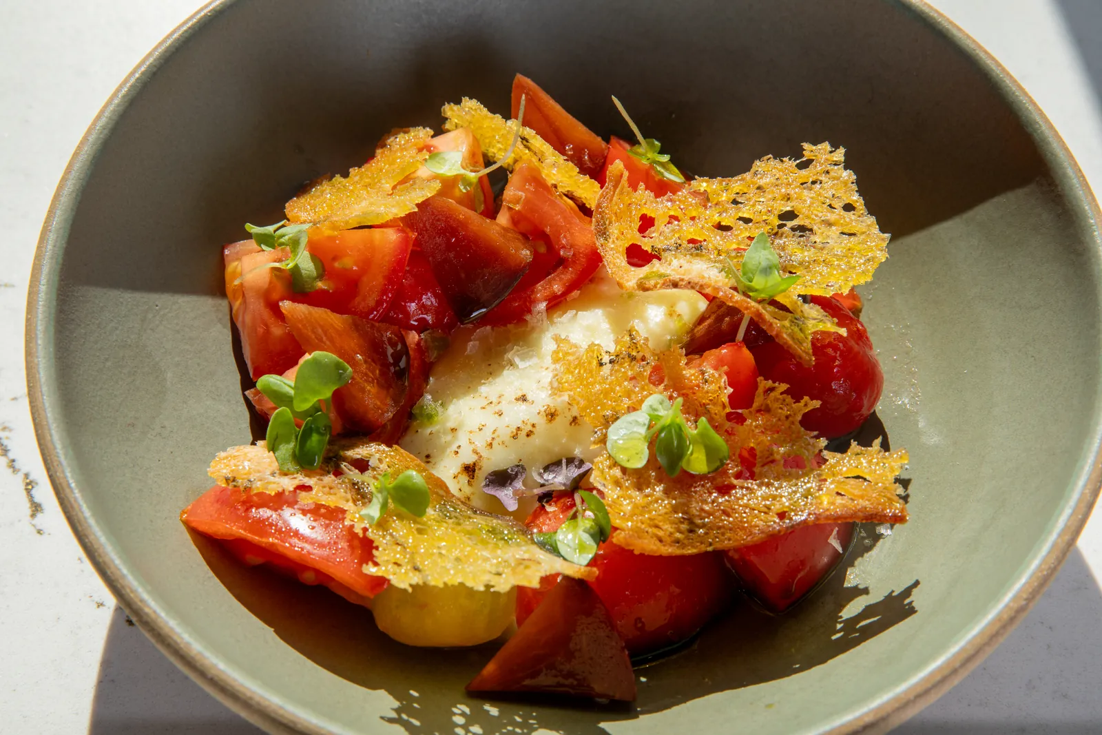 Red and yellow tomato wedges around soft white cheese under lacy golden crisps, in a sage-green bowl