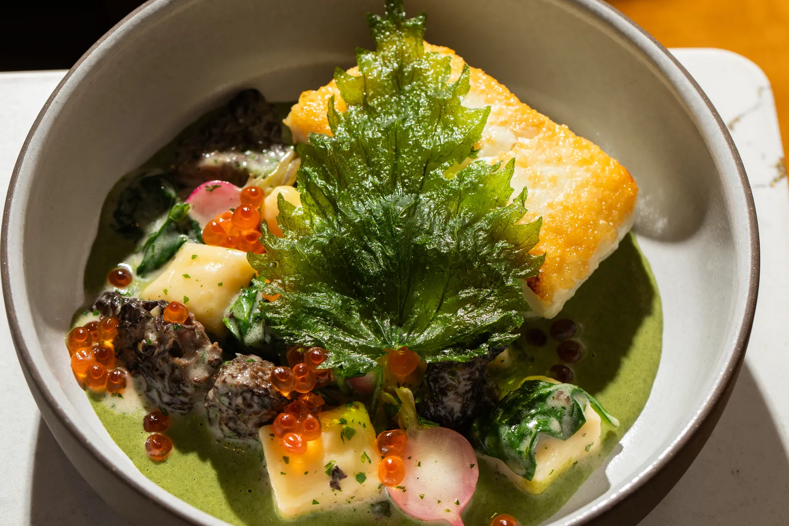 A seared white fish fillet with a crisp green leaf, orange roe, sliced radish and dark mushrooms in a green sauce, in a grey bowl