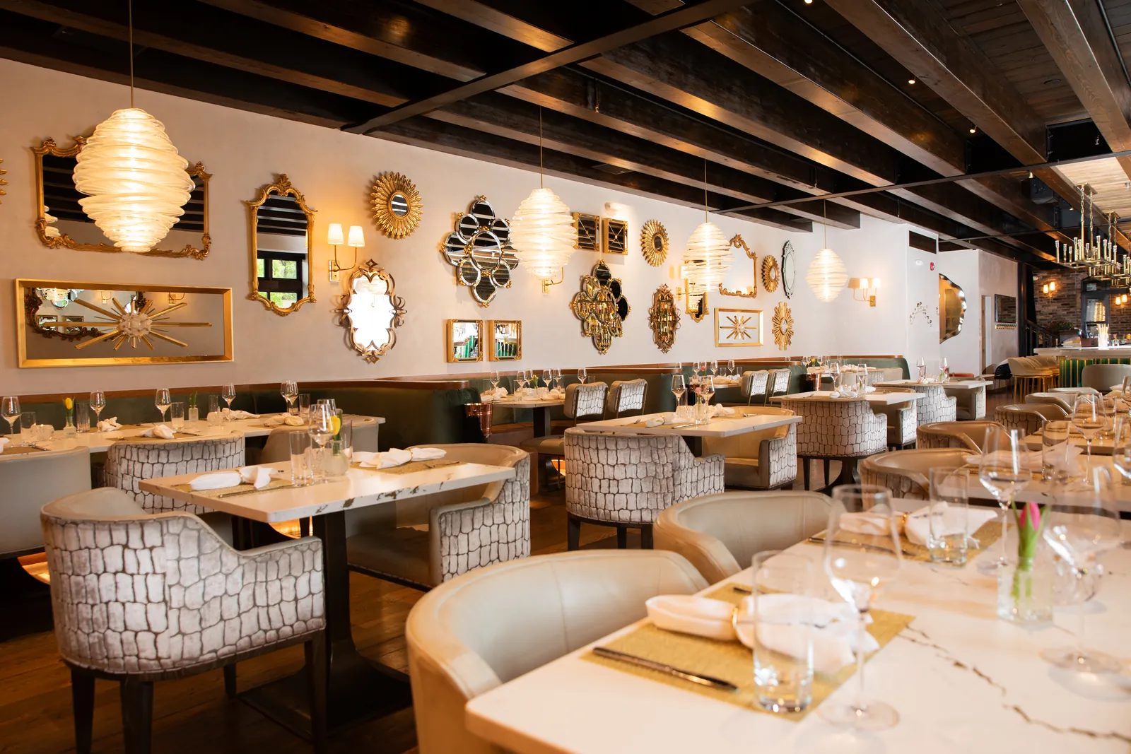 An empty dining room with gold-framed mirrors on a white wall, ribbed glass pendant lamps and tables set with glasses and napkins
