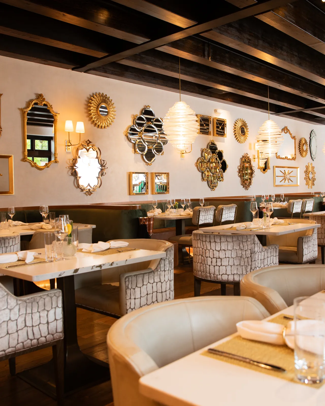 An empty dining room with gold-framed mirrors on a white wall, ribbed glass pendant lamps and tables set with glasses and napkins