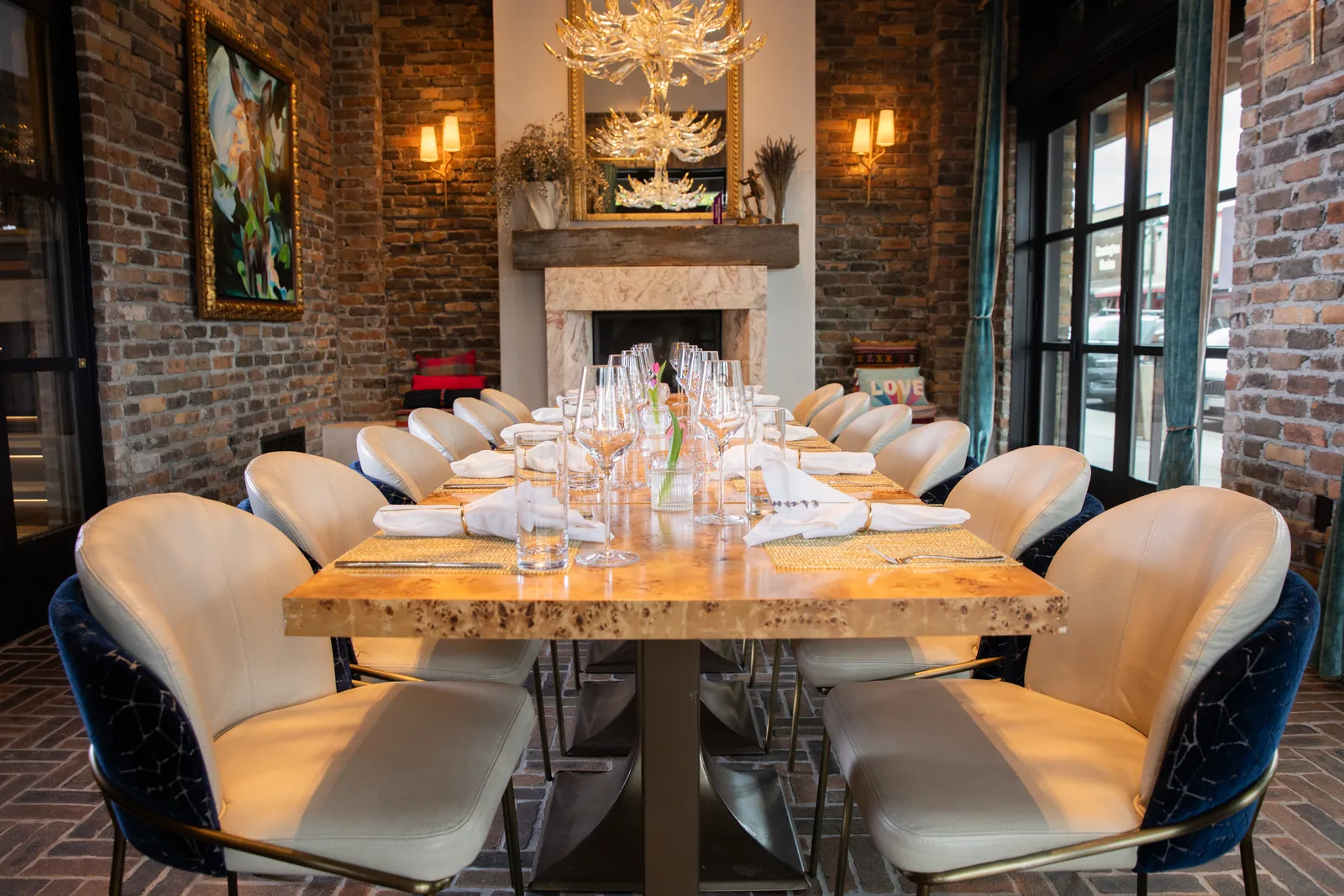 A long wooden table set with glasses and napkins between cream chairs in a brick room, a stone fireplace with a mirror and chandelier at the far end