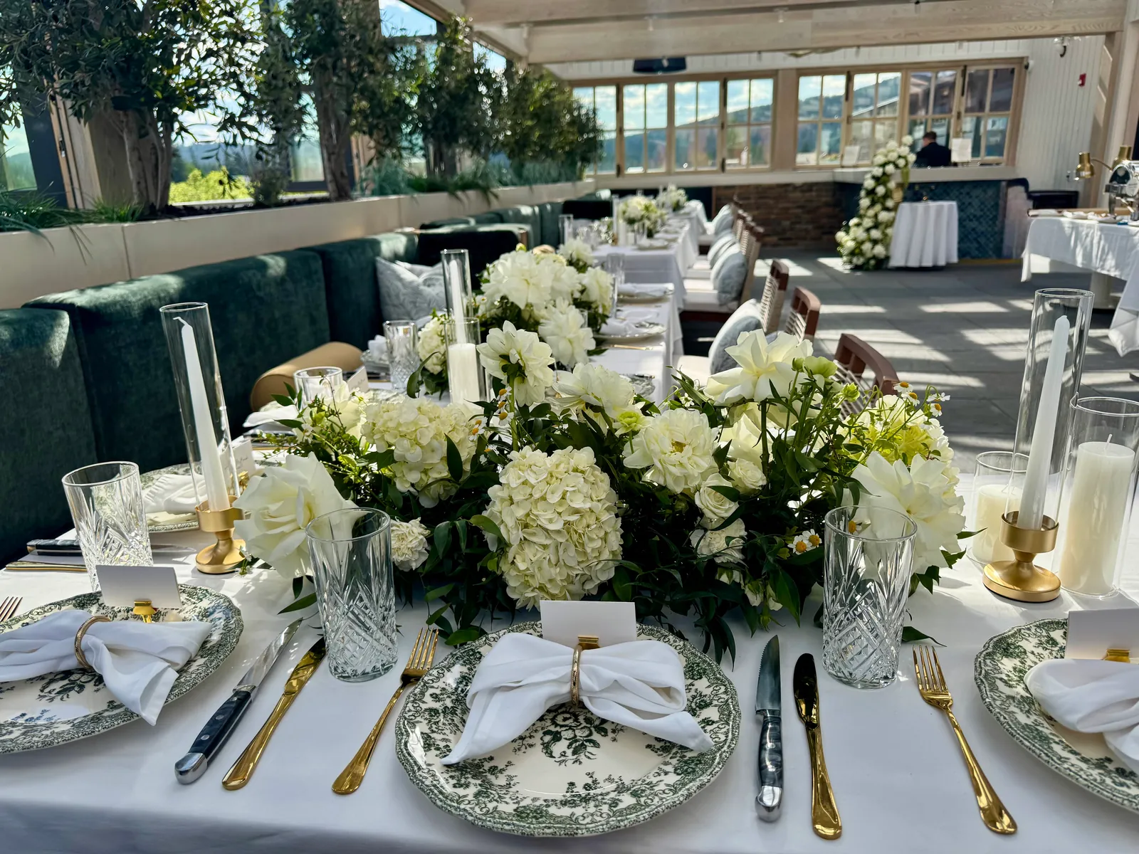 A long table in white linen with white hydrangeas and other white flowers, green-patterned plates, gold cutlery and taper candles, in a sunlit room with potted trees and windows onto hills